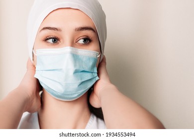 Nurse young medic standing near white wall and looking at the camera. Female doctor dressed in medical gown and surgical hat. Pandemic virus coronavirus concept, disposable face mask, copy space.
