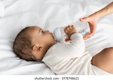 Nursery And Childcare Concept. Portrait Of Cute Little Black Kid Drinking Milk, Babysitter Holding Baby Bottle In Hands, Feeding Child Who Lying On White Blanket On The Bed In The Bedroom At Home