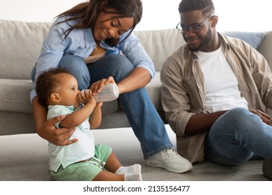 Nursery Concept. Smiling Loving African American Parents Feeding Their Baby Boy From Bottle, Cute Black Toddler Drinking Water Or Milk. Millennial Family Sitting On Couch, Spending Time With Child