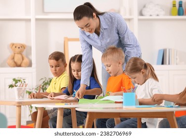 Nursery teacher with group of cute little children drawing and cutting paper at desks in kindergarten. Playtime activities
