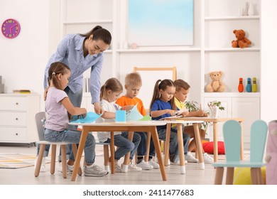 Nursery teacher with group of cute little children drawing and cutting paper at desks in kindergarten. Playtime activities - Powered by Shutterstock