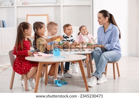 Nursery teacher and group of cute little children making toys from color paper at desks in kindergarten. Playtime activities