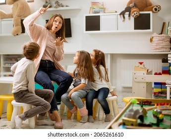 Nursery teacher talking story to group of children sitting on a small chairs at kindergarten - Shutterstock ID 2065261610