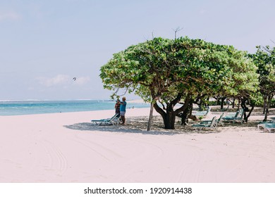 Nusa-Dua, Bali - 20.09.2019. White Sand Beach, Tourists Standing Next To Sun Lounger Under Tree And Photographing Something. Against The Backdrop Of The Ocean, Sunny Day, Vacation.