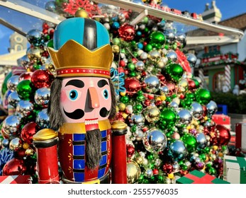 A Nutcracker puppet soldier stands in front of a Christmas tree decorated with colorful balls.