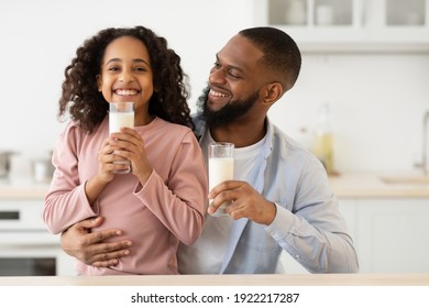 Nutrition Concept. Portrait Of Smiling Black Girl Holding Glass And Drinking Milk With Her Father In The Kitchen. Happy Dad Hugging His Cute Curly Daughter, Having Healthy Meal Together