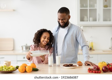 Nutrition Concept. Portrait Of Smiling Black Girl Pouring Fresh Milk From Bottle In Glasses Standing With Her Father In The Kitchen. Happy Dad Hugging His Curly Daughter, Having Healthy Meal Together