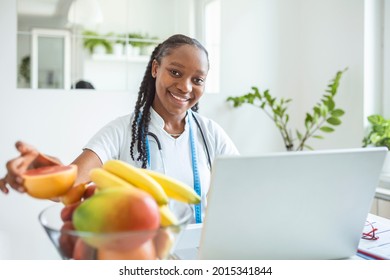 Nutritionist Doctor With Fruits In Office. Nutritionist Working On Laptop And Writing Diet Plan For Patient. Portrait Of Female Nutritionist In Her Office. Smiling Young Dietician Sitting At Desk