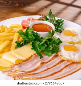 Nutritious breakfast combination includes fried eggs with bacon slices and cheese. Hot food is served beside french fries and red tomato sauce.