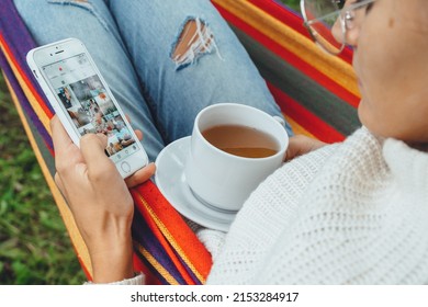 NY, USA - 2 May,2022- Millennial Woman Using Smart Phone Relaxing On Hammock Chatting Using Smartphone Application Check Social Media Network Scrolling Instagram Like Photos Drinking Tea Outdoor.