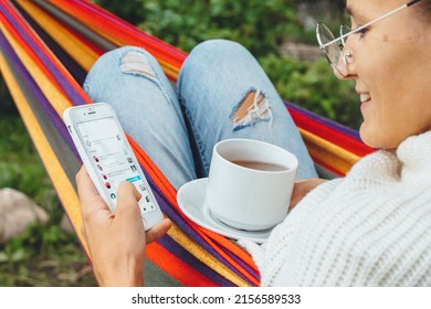 NY, USA - 2 May,2022- Millennial Woman Using Smart Phone Relaxing On Hammock Chatting Using Smartphone Application Check Social Media Network Scrolling Instagram Like Photos Drinking Tea Outdoor.