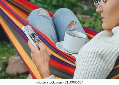 NY, USA - 2 May,2022-millennial Woman Using Smart Phone Relaxing On Hammock Chatting Using Smartphone Application Check Social Media Network Scrolling Instagram Like Photos Drinking Tea Outdoor.