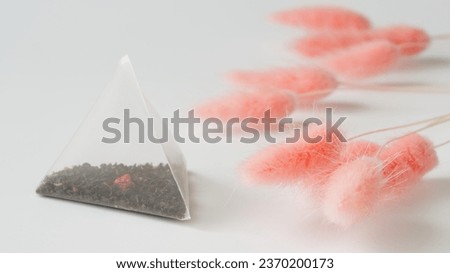 Nylon white pyramid tea bag with black tea, with the addition of fruits and berries, lies on a white background next to pink dried flowers. Flavored tea in triangular bags. Photo. Close-up