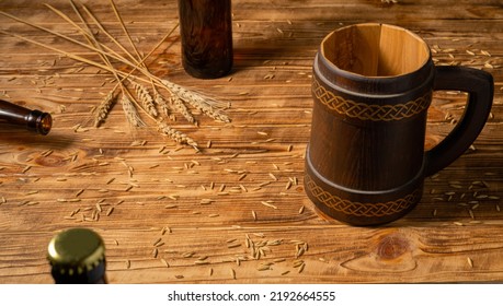 Oak Mug With A Handle And A Glass Brown Bottle On A Wooden Table In A Pub Or Bar. Handmade Wooden Tankard For Beer Or Kvass. Brown Mug With Ornament, Wheat Grains And Spikelet On A Wooden Surface.