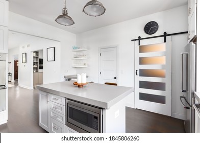 OAK PARK, IL, USA - AUGUST 7, 2020: A White Kitchen With Stainless Steel Appliances, Grey Countertops, And Hardwood Floors Through The House. A Barn Door Hangs With Light Shining Through.