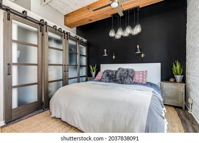 OAK PARK, IL, USA - NOVEMBER 2, 2020: A Bedroom In A Loft Condo With An Exposed Wood Ceiling, Glass Sliding Doors To A Closet, Black Accent Wall, And A Fancy Glass Light Hanging Above.