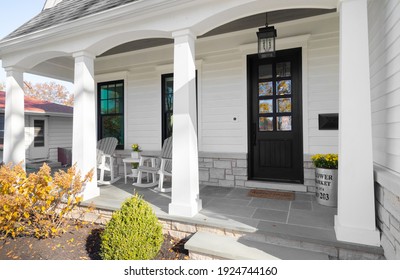 OAK PARK, IL, USA - OCTOBER 28, 2020: A New, White Modern Farmhouse With A Dark Wood Door With Windows, White Pillars, A Stone Floor, And Patio Furniture.