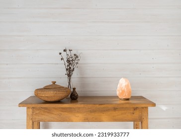Oak side table with rustic accessories against white painted wood panelled wall including salt lamp, basket and dried seed pods (selective focus)