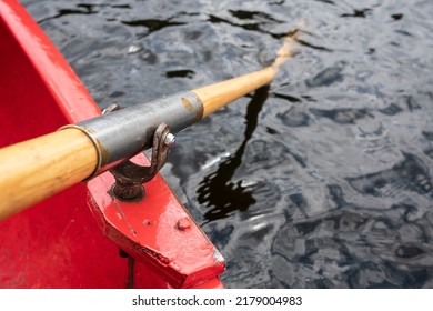 Oar Is Held On An Oarlock Attached To A Red Boat, Against The Background Of Water. 