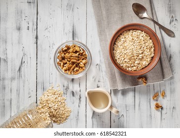 Oat Flakes In Brown Clay Bowl Ready To Cook, Scattered Oat Flakes, Walnuts, Jug With Milk And Spoon On The Light Wooden Table. View From Above