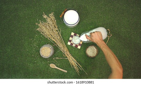 Oat Milk And Oats On The Lawn.  Top View. 
The Male Hand Takes A Jug And Pours Milk Into A Mug. 
