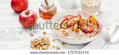 Oat porridge with caramelized apples and nuts on white wooden background.