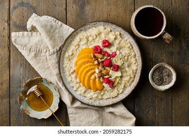 Oat Porridge With Sliced Peach, Fresh Raspberries, Chopped Almond Nuts, Coconut Chips, Chia Seeds, Honey And Tea. Classic Bunting Breakfast Served In Ceramic Bowl On Wooden Background