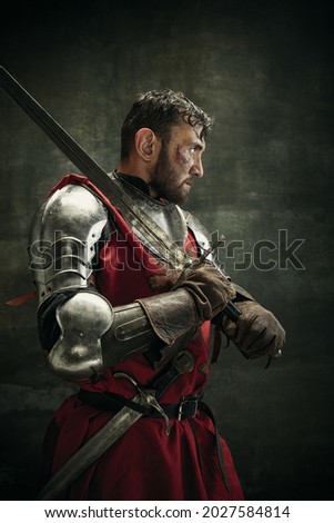 Oath to the king. Portrait of one brutal bearded man, medeival warrior or knight with dirty wounded face holding sword and shield isolated over dark background.
