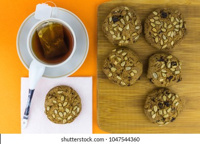 Oatmeal Cookies With Raisins And Sunflower Seeds On A Bamboo Board, On The Left Tea With Tea Leaves With A Bag In A Mug, One Cookie On A Napkin Next To A Cup, Top View, On An Orange Background