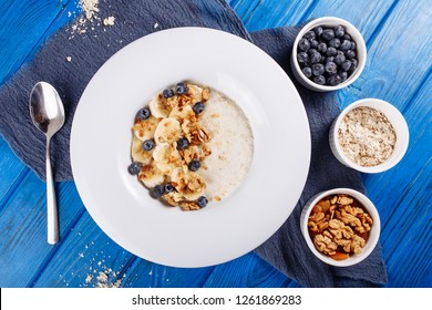 Oatmeal Muesli Blueberry And Banana Top Down View. Porridge Bowl With Walnut And Berry Ingridient Healthy Food Breakfast On Wooden Blue Background. Cereal Flakes Of Milk Or Yogurt In White Plate