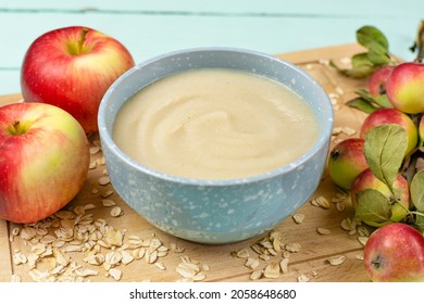 Oatmeal Porridge For The Baby From Ground Cereals In A Blue Bowl, Red Ripe Apples On A Wooden Board On A Blue Background Close-up. Baby Nutrition, The First Complementary Feeding Of A Child.