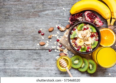 Oatmeal Porridge With Banana, Kiwi Fruit, Pomegranate, Nuts And Honey In A Bowl With Egg And Fresh Orange Juice For Healthy Breakfast On Rustic Wooden Background. Top View