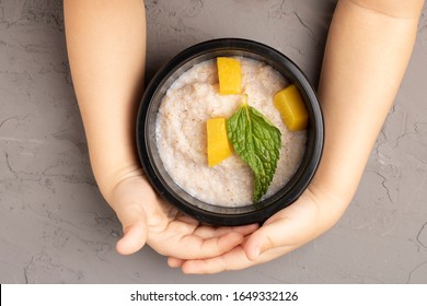 Oatmeal Porridge Bowl With Fruits In Hands. Hands Holding Bowl Of Oat Porridge With Ananas Pieces And Mint Leaf. Top View Food