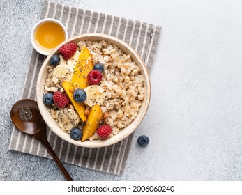 Oatmeal porridge in ceramic bowl decorated with fresh berries, banana slices, nectarine pieces and chia seeds served with honey. Healthy breakfast concept. White table top view. Copy space.