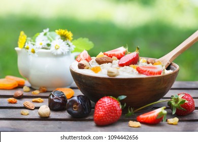 Oatmeal Porridge In Wooden Bowl With Fresh Strawberries, Mix Nuts And Dried Fruit. Outdoor Image, Green Meadow In Background. Decorated With Wild Fowers In A Small Vase. 