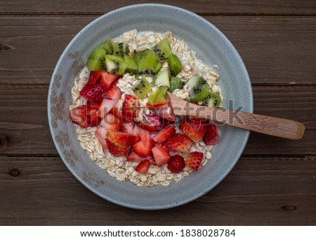 Oatmeal in a tan plate decorated with diced strawberries and kiwi on wooden table, whole plate, centered, top view, with silverware spoon