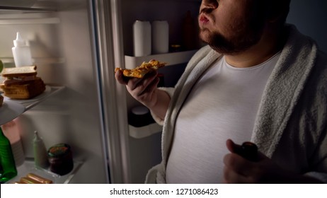 Obese bachelor eating pizza near fridge, holding beer, unhealthy lifestyle