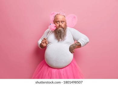 Obese bearded man dresses for hallooween party, plays fairy with magic wand, looks surprisingly at camera, wears wings and crown on bald head, being at childrens holiday. Funny dad princess.