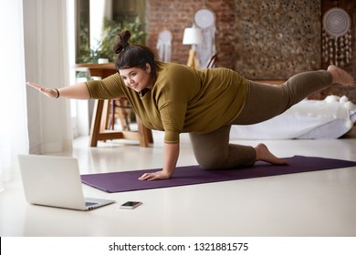 Obese Chubby Young European Female With Hair Knot Practicing Yoga Or Pilates Indoors On Mat, Doing Exercises To Strengthen Core, Watching Video Lesson Online In Front Of Open Laptop Computer On Floor