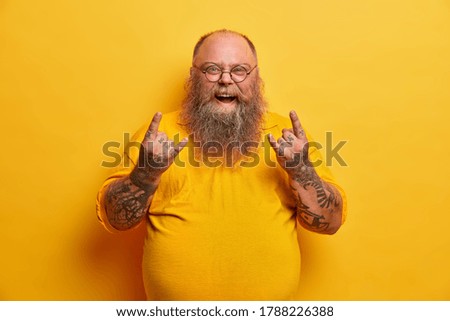 Obese funny man in yellow t shirt, shows heavy metal sign, attends concert of favorite music band, has big belly, tattooed arms and beard, wears round glasses. Overweight rock fan gestures indoor
