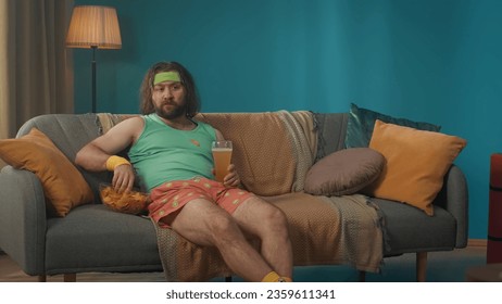 Obese middle-aged man with long hair sits in a room on a couch carelessly eating chips and drinking beer. The man is carefully watching a TV program on the television. - Powered by Shutterstock