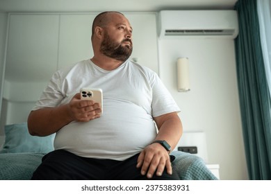 Obese unshaved man with phone and fitness tracker looking at window. Overweight middle-aged man thinks on weight reduction, diet and calories calculation. Weight control and healthy food concept - Powered by Shutterstock