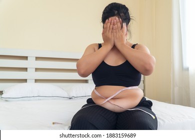 Obesity unhealthy concept. Overweight Asian woman sitting on white bed while holding hands cover on her face at home. Upset female measure tape on body suffering from extra weight in the bedroom.