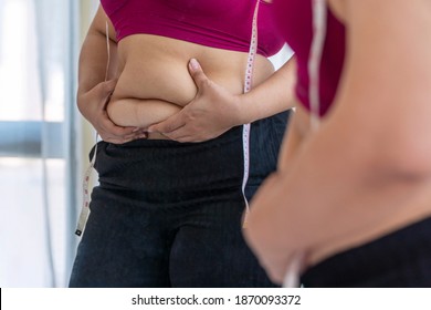 Obesity unhealthy concept. Reflection of overweight woman wear sport bar with measure tape on body pinches fat on her belly large size while standing front mirror. select focus