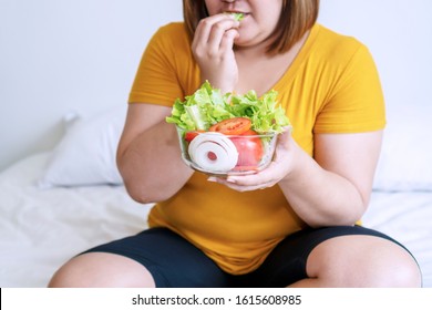 Obesity Young Women Eat Green Organic Vegetables While Sitting On The White Bed. Fat Female Holding Bowl Salad Vegetarian While Eating Lettuce In Her Hand. Concept Food Good Healthy For Weight Loss.