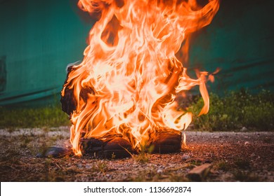 Object seen burning from the front on gravel floor and green background. Black and worn motorcycle boots burning and are on fire. Motorcycle shoes on fire. Concept of burning shoes.