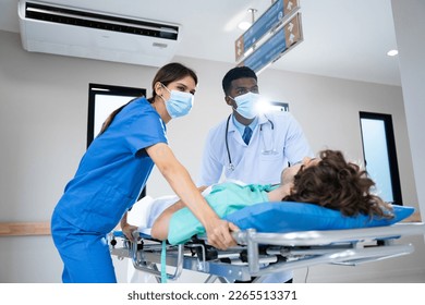 Oblique and low angle view of young African male medicine doctor and young Caucasian female nurse in face masks, urgently pushing Caucasian male patient on emergency stretcher bed at hospital corridor - Powered by Shutterstock