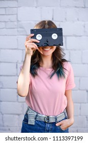 obscured view of smiling woman covering eyes with retro video cassette in hand against white brick wall