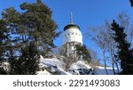 An observation tower in Mikkeli. The white tower on top of hill which is photographed from below. Clear blue sky and snow is in a picture. Some trees are near the tower. A sunny winter day.