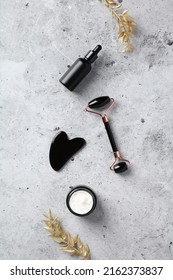 Obsidian stone face roller, gua sha, serum dropper bottle, moisturizer cream jar and golden branches on concrete background. Face skin treatment, anti-aging concept. - Shutterstock ID 2162373837